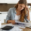 A woman contemplates her bills while sitting at her kitchen table.